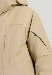 Beige vadderad jacka med huva, med en strukturerad design, sömdetaljer och en sidoficka med svart accent. Slät textur.