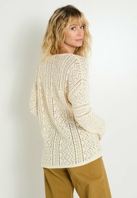 Mujer rubia con cabello ondulado lleva un suéter de punto de color crema y con estampados, y pantalones marrones, mirando hacia atrás por encima del hombro frente a un fondo blanco.