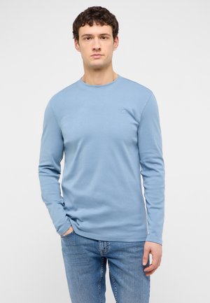 Young man with curly dark hair wearing a light blue long-sleeve shirt and blue jeans, standing with one hand in pocket.