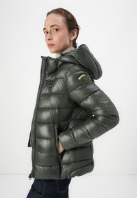 Veste rembourrée verte avec une finition brillante, fermeture éclair à l'avant, capuche et poches latérales. Présente un patch logo sur la manche gauche.