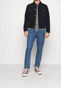 Mann in dunkelblauer Jacke, grauem Hemd, blauen Jeans und braunen High-Top-Sneakern, der vor einem schlichten hellen Hintergrund steht.