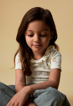 Młoda dziewczyna o długich brązowych włosach, ubrana w biały kwiatowy, falbaniasty top z krótkim rękawem i niebieskie dżinsy, siedząca na beżowym tle.