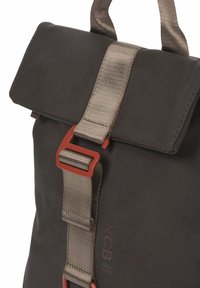 Zwarte stoffen rugzak met een overslagklep, beige riem en rode gespsluiting. Inclusief logo detail aan de zijkant.