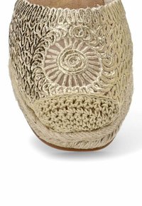 Zapato de punto beige con un patrón circular metálico y puntera texturizada. Presenta un frente de ganchillo y detalles trenzados a lo largo de la suela.