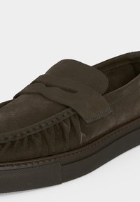Σκούρο καφέ μοκασίνι από σουέτ με ραφές μοκασίνου, λεπτομέρεια με λουράκι «penny» και μαύρη ελαστική σόλα.
