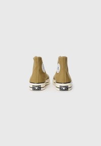Bruine hoge sneakers van canvasmateriaal, met een witte strook, zwarte zool en cirkelvormig logo-detail aan de buitenkant. Verhoogd hakontwerp.