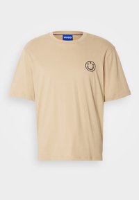 T-shirt en coton beige à manches courtes, col rond, avec un logo smiley noir et le texte "HUGO" en dessous sur le côté gauche de la poitrine.