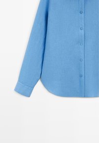 Lyseblå langærmet skjorte med knapper, buet skørt og knappede manchetter, vist på en ensfarvet hvid baggrund.