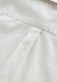 Lyst beige stof med en glat tekstur, fremhævet af et plisseelement og synlige sømme langs den øverste kant.