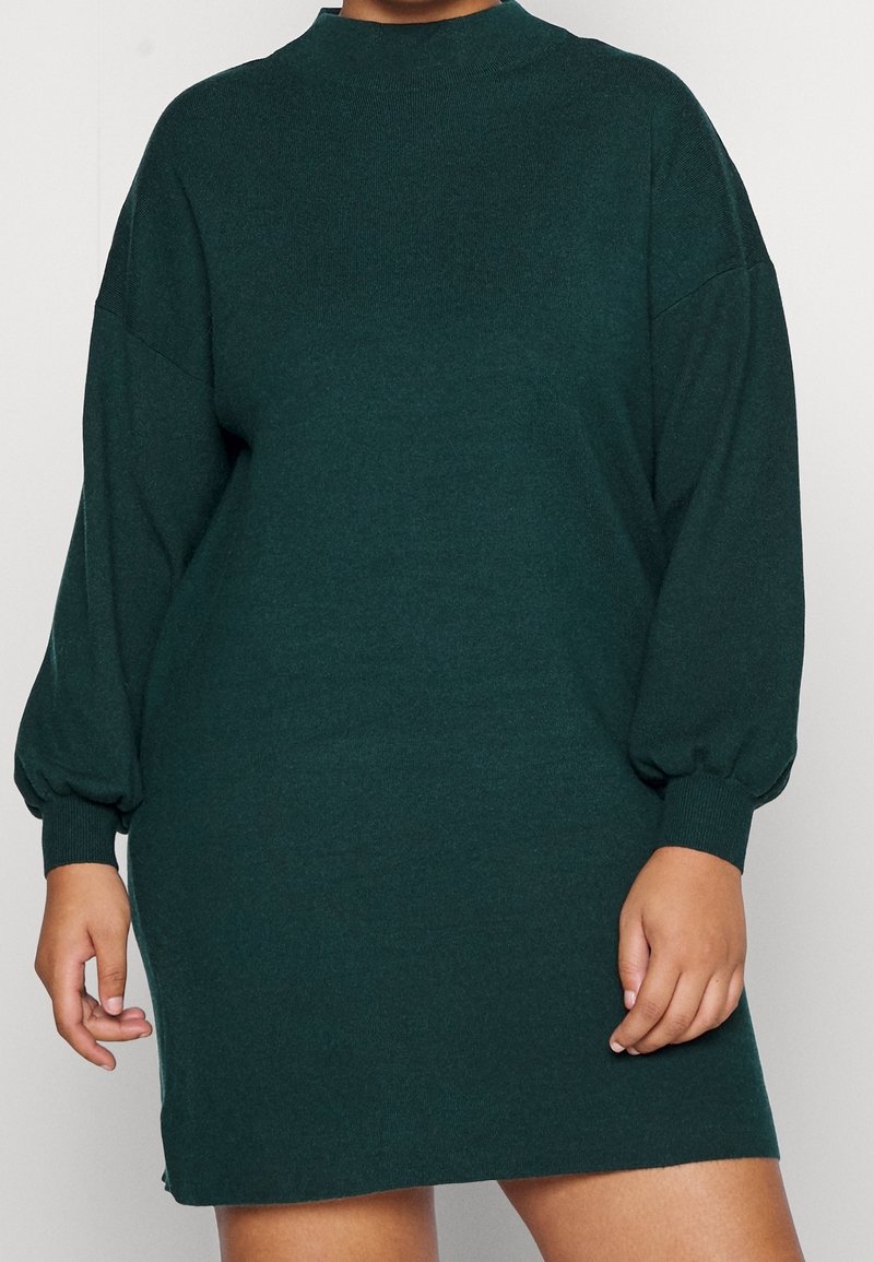 Robe pull verte à manches longues avec col montant et poignets froncés. Texture douce, coupe ample et longueur jusqu'aux genoux avec fentes latérales.