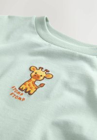 Hellgrünes Baumwoll-T-Shirt mit einem aufgenähten Giraffendesign in Gelb und Braun, darunter der Text "STOMP STOMP" in Orange.