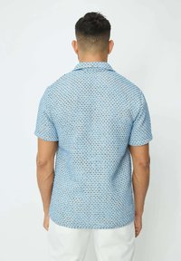 Chemise à manches courtes de couleur bleu clair, avec un motif texturé comportant de petites formes géométriques. Design avec col et coupe droite.