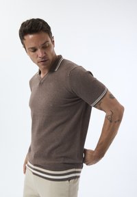 Polo shirt marron à manches courtes en tissu texturé, col et ourlet rayés blancs, porté avec un pantalon de couleur claire.