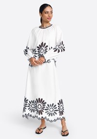 Hvid langærmet kjole med sort blomsterbroderi langs ærmerne og kanten, med samlet halsudskæring og elastiske manchetter. Flade sandaler.