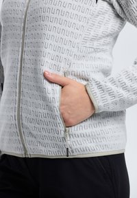 Chaqueta de forro polar en gris claro con un sutil patrón de texto negro, que cuenta con un cierre frontal con cremallera, un bolsillo lateral y puños elásticos.