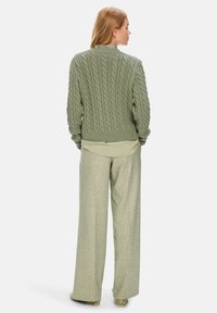 Grüner Kabelstrickpullover mit geripptem Saum, über einem hellgrünen Shirt getragen, kombiniert mit weiten, strukturierten Hosen in einem passenden Farbton.