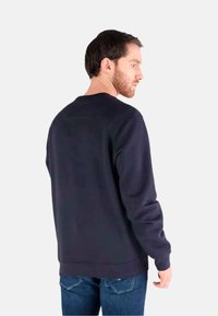 Hombre con cabello corto y oscuro y barba, vistiendo una sudadera azul marino lisa y vaqueros azules, de perfil sobre un fondo blanco.