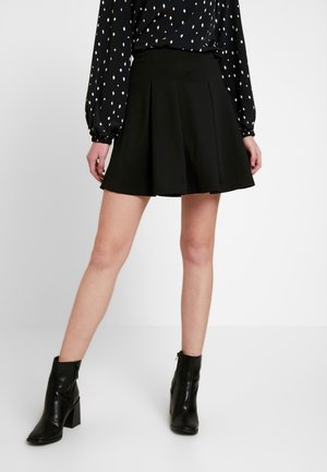 Kvinde iført en sort plisseret nederdel, sorte ankelstøvler med blokhæle og en sort langærmet top med hvide diamantmønstre.