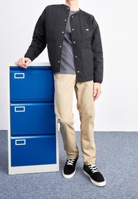 Person wearing black jacket, gray shirt, beige pants, and black sneakers stands next to a blue three-drawer filing cabinet on a gray carpet.