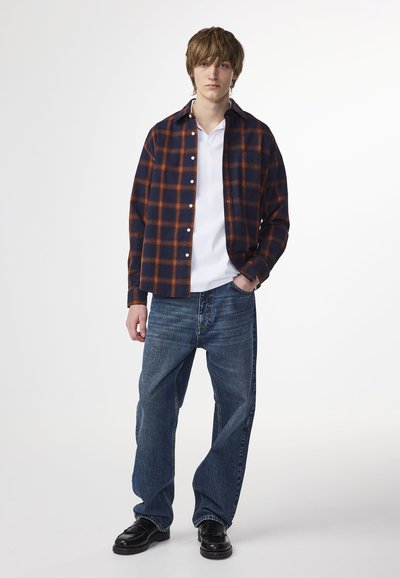Flanellhemd in marineblau und orange kariert, mit Knopfverschluss. Darunter ein weißes T-Shirt, kombiniert mit locker sitzenden blauen Jeans und schwarzen Schuhen.