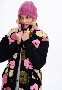 Fleecová bunda s černým základem a velkými květinovými vzory v růžové, krémové a zelené barvě. Vysoký límec a přední kapsy, doplněné růžovou žebrovanou čepicí.