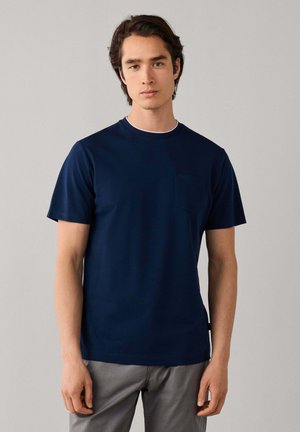 Jonge man met bruin haar, die een marineblauw T-shirt met korte mouwen en een grijze broek draagt, staand tegen een effen lichtgrijze achtergrond.