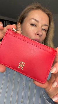 Portacarte in pelle rossa con chiusura a zip, dettaglio logo dorato e bordi cuciti, tenuto contro uno sfondo di camicia a righe.