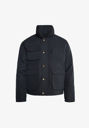 Granatowa pikowana kurtka z wysokim kołnierzem, wieloma przednimi kieszeniami, mosiężnymi zatrzaskami oraz kieszenią zapinaną na zamek błyskawiczny na klatce piersiowej.