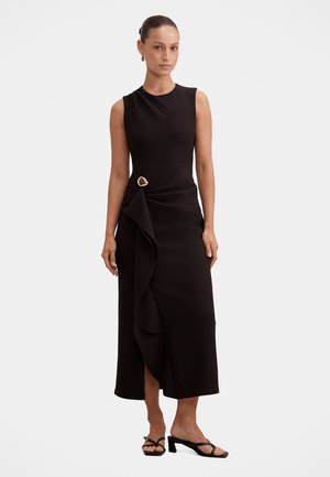 Femme aux cheveux attachés portant une robe midi noire sans manches avec un volant latéral et une broche dorée, assortie à des sandales à talons ouvertes noires.