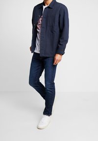 Chaqueta azul marino con cuello, cierre de botones y dos bolsillos frontales, combinada con jeans oscuros y zapatillas blancas.