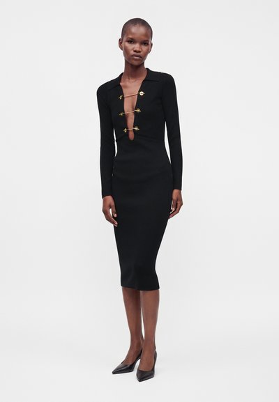 Femme grande portant une robe midi noire ajustée à manches longues, fermée par trois épingles de sûreté dorées sur un profond décolleté en V, associée à des talons noirs.