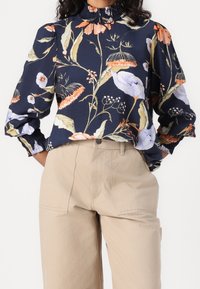 Navy floral blouse with long, puffed sleeves and a ruffled collar. Features colorful flowers and greenery, paired with beige trousers.