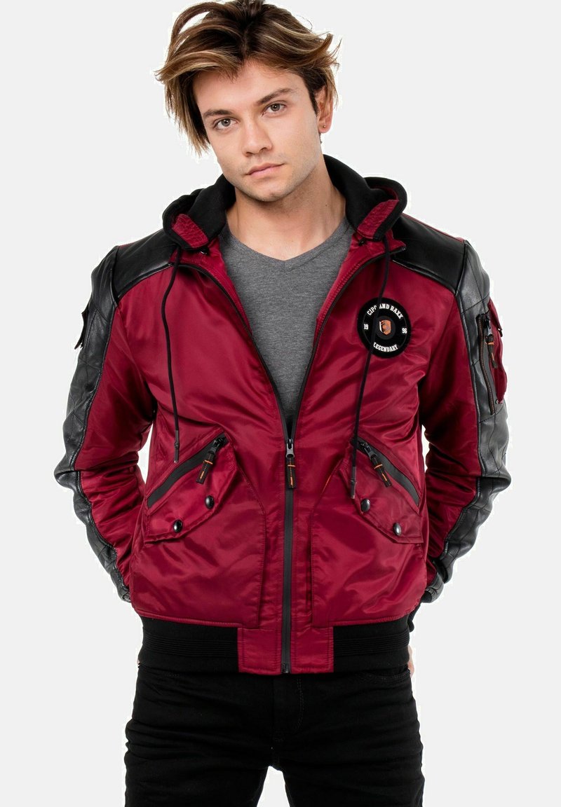 Rote und schwarze Bomberjacke mit einem Reißverschluss vorne, zwei Fronttaschen und einem runden Logo-Patch auf der linken Seite. Glatte Oberfläche und taillierte Passform.