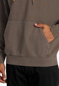 Bruine sweatshirt met een voorzak, capuchon met trekkoord en geribbelde manchetten. Zachte stof; comfortabele pasvorm. Gecombineerd met zwarte broek.
