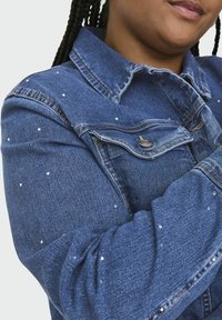 Denim jakna s klasičnim ovratnikom, svetlo modre barve, s majhnimi kovinskimi okrasnimi elementi na rokavih, z sprednjim žepom in detajli na gumbih.