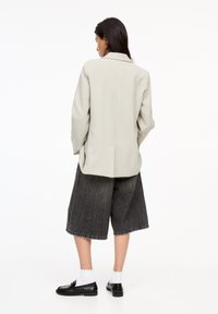 Hellgrauer oversized Blazer mit glatter Textur, kombiniert mit lockeren dunklen Denim-Shorts, schwarzen Slippern und weißen Knöchelsocken.
