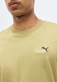 Homme portant un T-shirt Puma vert clair avec un logo Puma noir et un texte blanc sur le côté gauche de la poitrine sur un fond uni.