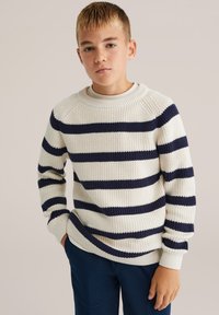 Sweter robiony na drutach w kolorze kości słoniowej z granatowymi poziomymi paskami, okrągły dekolt, ściągacze na rękawach i u dołu. Połączony z ciemnoniebieskimi spodniami.
