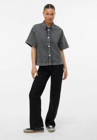 Camicia nera in denim a maniche corte con colletto, caratterizzata da bottoni grandi e una vestibilità rilassata, abbinata a pantaloni neri e sneakers.