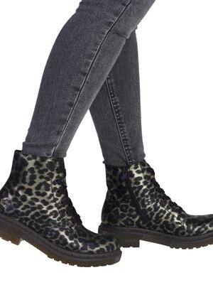 Jambes en jean skinny gris foncé portant des bottes à lacets noir et or à imprimé léopard avec fermetures éclair latérales, debout sur un fond blanc.