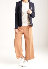 Nainen, joka on pukeutunut tummansiniseen bleiseriin, valkoiseen neulepaitaan, kamelinvärisiin leveälahkeisiin housuihin ja valkoisiin lenkkareihin, seisoo tasaisella taustalla.