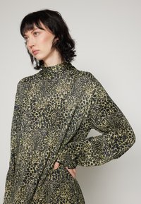 Blouse à imprimé animal avec motifs verts et noirs, col haut, manches longues et tissu texturé avec une coupe décontractée.