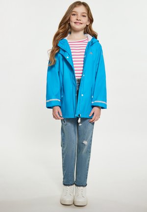 Giacca impermeabile blu brillante con cappuccio, caratterizzata da rifiniture bianche. Abbinata a una maglietta a righe rosse e bianche e a jeans chiari sdruciti.