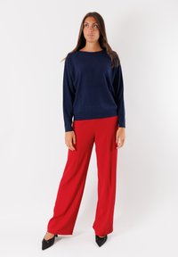 Maglione lavorato a maglia di colore blu navy con scollatura ampia, abbinato a pantaloni a gamba larga rossi e décolleté neri a punta. Texture liscia, silhouette moderna.