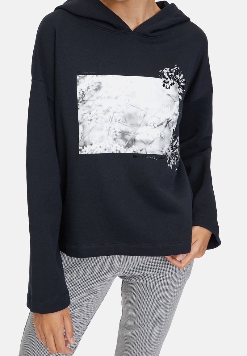 Personne portant un sweat à capuche noir avec un motif floral blanc et un pantalon à carreaux gris clair.