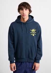 Sweatshirt cu glugă de culoare albastru închis, cu buzunar frontal, având o grafică galbenă cu cartofi prăjiți și raze pe partea stângă a pieptului. Textură din amestec de bumbac.