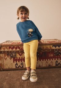 Blå sweatshirt med sol- och stjärndesign, gula leggings med stjärnapplikationer och guldglittriga skor. Stylad mot en texturerad bakgrund.