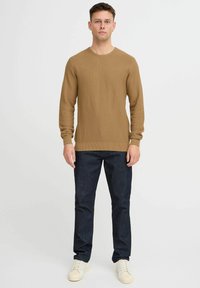 Suéter marrón de punto con cuello redondo, combinado con jeans azul oscuro y zapatillas crema. El suéter presenta un patrón texturizado y puños acanalados.