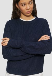 Pull en maille bleu marine avec un col rond, un tissu texturé et des manches longues légèrement oversize. Superposé sur un haut blanc.