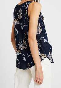 Haut sans manches floral bleu marine en tissu léger, avec des fleurs bleu clair et beige, dont l'ourlet arrière est plus long que l'avant.